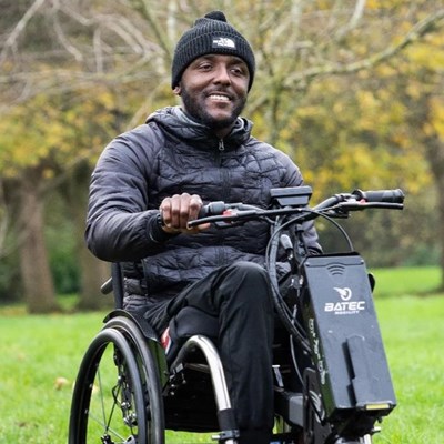 Image of a gentleman smiling while he wheels in a powered wheelchair. There a big green trees behind him. 