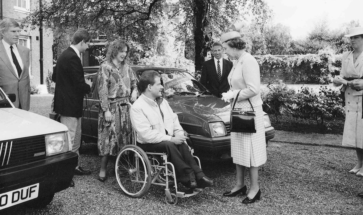 Her Majesty The Queen standing in front of a car and talking to a gentleman who is sitting in a wheelchair. They are both smiling.