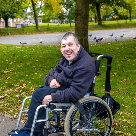 Kevin is outside in a park in the autumn, sat comfortably in his wheelchair as he smiles to camera 