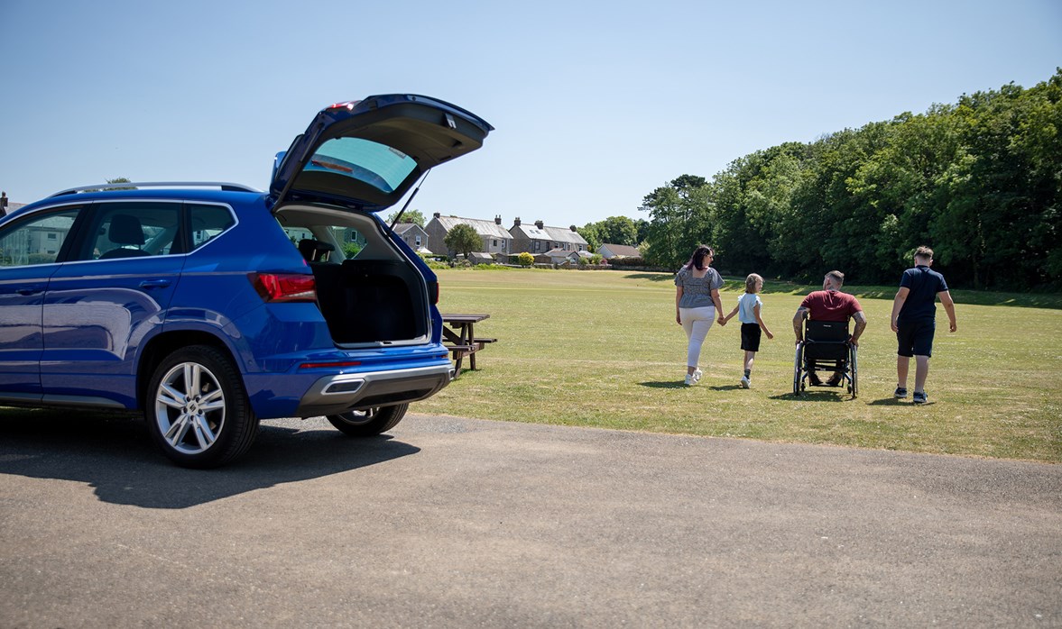 A blue car sits near a park with the boot open. A family walk away in the distance.
