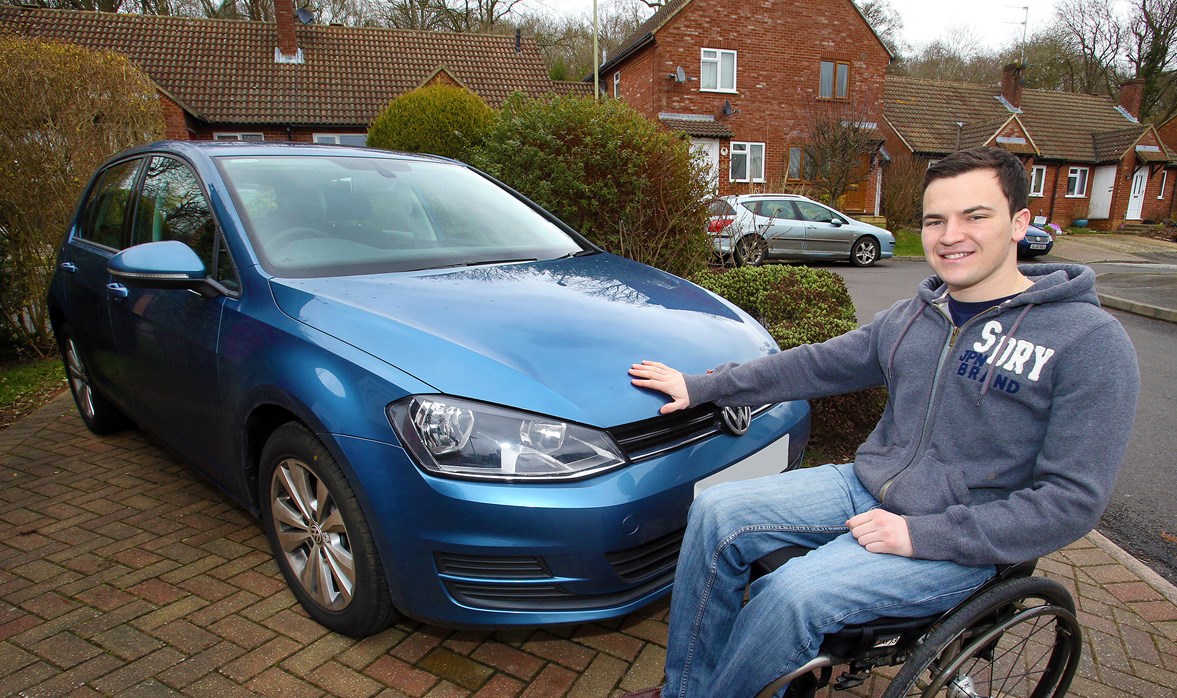Aaron is sitting in his wheelchair, with his right hand on the front bonnet of his specially adapted car