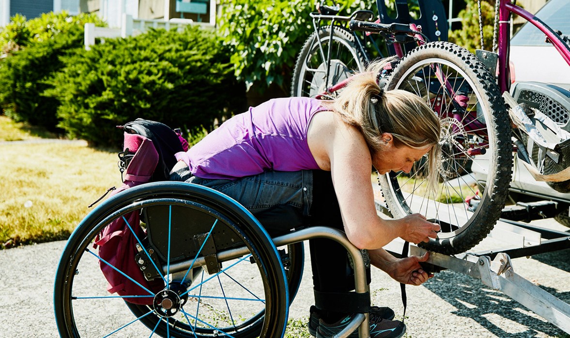 A lady in a wheelchair wearing a purple top is attaching a bike to the boot of a car.