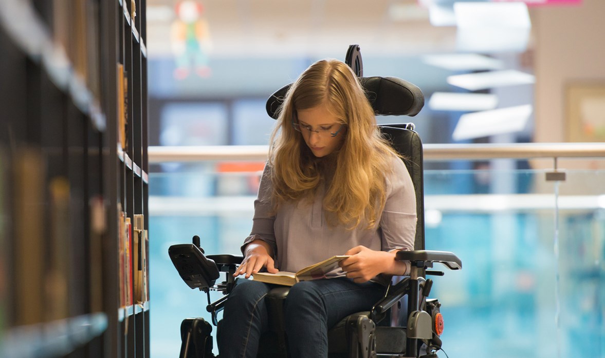 A young lady sitting in a wheelchair whilst next to a book shelf in a library is reading a book which is placed on her lap. 