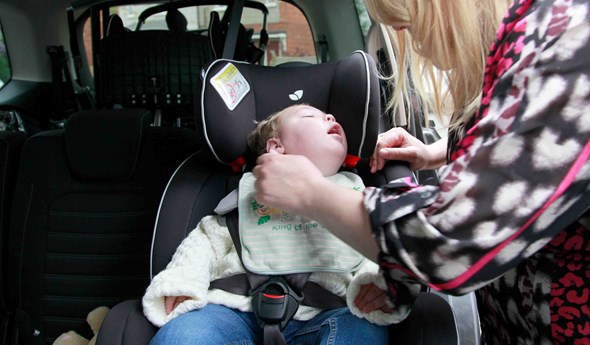A baby boy is within a car, sitting in a car seat while his mum is securing the car seat safely. 