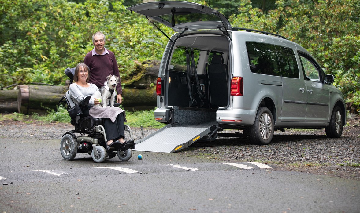 Michelle is outside her WAV in her motorised wheelchair, with her dog on her lap and her husband stood beside her.  