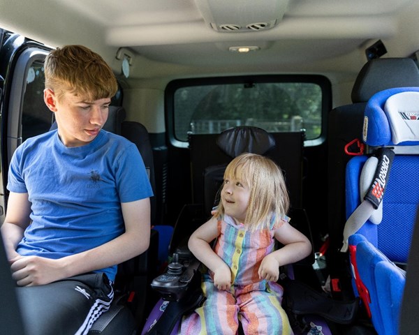 Poppy is sitting in her powered wheelchair in her vehicle. To the left of her is her old brother Liam, Poppy and Liam and smiling at one another. To the right of her is her car seat.