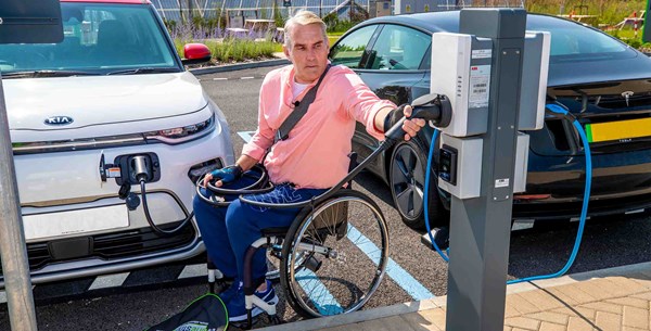 A man in a wheelchair is plugging in his electric car into an electric vehicle charging-point. 