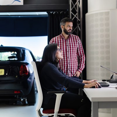 A woman and a man look at a computer screen with a car in the background